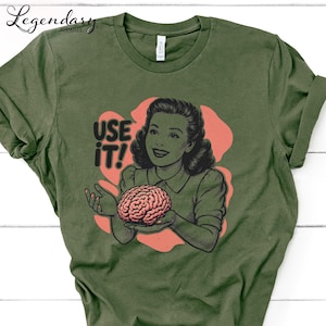 Pu&ograve; includere: T-shirt verde oliva con un'illustrazione retr&ograve; di una donna che tiene in mano un cervello. L'immagine &egrave; ambientata su uno sfondo color corallo con le parole "USE IT!" in grassetto. La maglietta ha un classico girocollo.