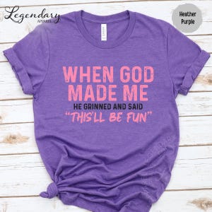 A purple t-shirt with a humorous text printed on it, which reads "WHEN GOD MADE ME HE GRINNED AND SAID 'THIS'LL BE FUN'".