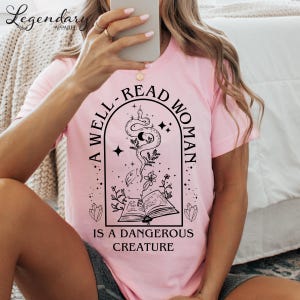 Może przedstawiać: Różowy T-shirt z czarnym nadrukiem graficznym, na którym widnieje napis "Dobrze czytająca kobieta jest niebezpiecznym stworzeniem". Grafika przedstawia węża owiniętego wokół książki z gwiazdami i kwiatami.