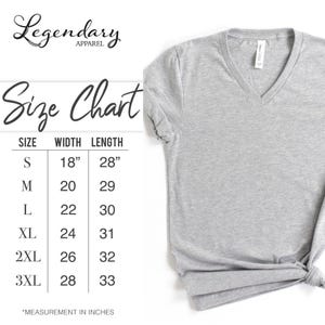 Pode incluir: Uma camiseta cinza claro com decote em V com uma tabela de tamanhos. A tabela de tamanhos mostra as medidas em polegadas para os tamanhos S a 3XL. A camiseta &eacute; da Legendary Apparel.