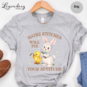 A grey t-shirt with a cartoon rabbit and a yellow duck holding a knife, accompanied by the text "MAYBE STITCHS WILL FIX YOUR ATTITUDE".
