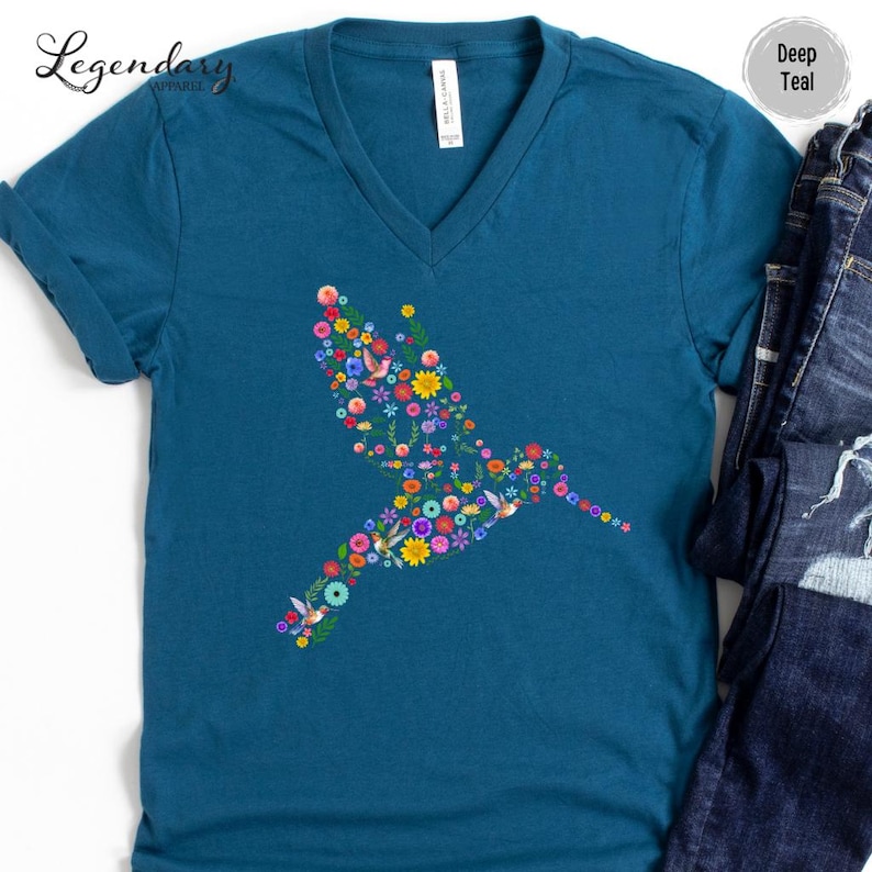 May include: A teal blue v-neck t-shirt with a hummingbird design made of colorful flowers. The hummingbird is facing right and is in flight.