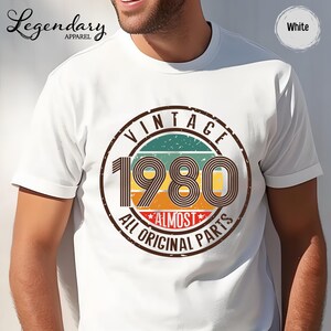 May include: White t-shirt with a vintage-style graphic. The graphic features the year "1980" in large numbers, surrounded by a circular design with the words "VINTAGE" and "ALL ORIGINAL PARTS". The shirt has short sleeves.