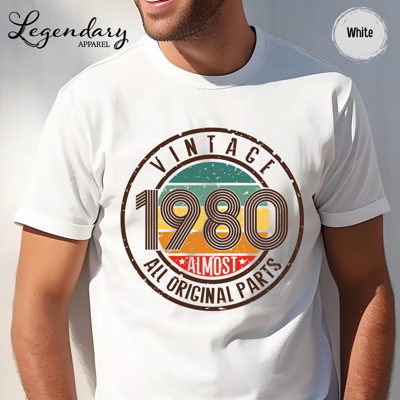 May include: White t-shirt with a vintage-style graphic. The graphic features the year "1980" in large numbers, surrounded by a circular design with the words "VINTAGE" and "ALL ORIGINAL PARTS". The shirt has short sleeves.