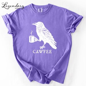 A purple t-shirt with a white illustration of a bird holding a cup of coffee, accompanied by the text "CAWFREE".