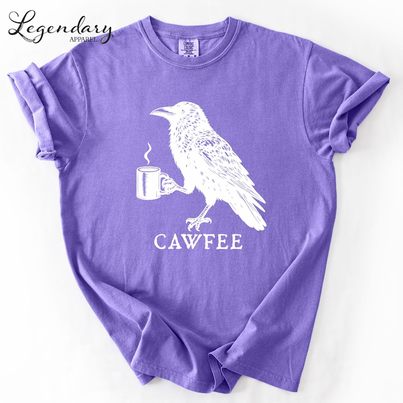 A purple t-shirt with a white illustration of a bird holding a cup of coffee, accompanied by the text "CAWFREE".