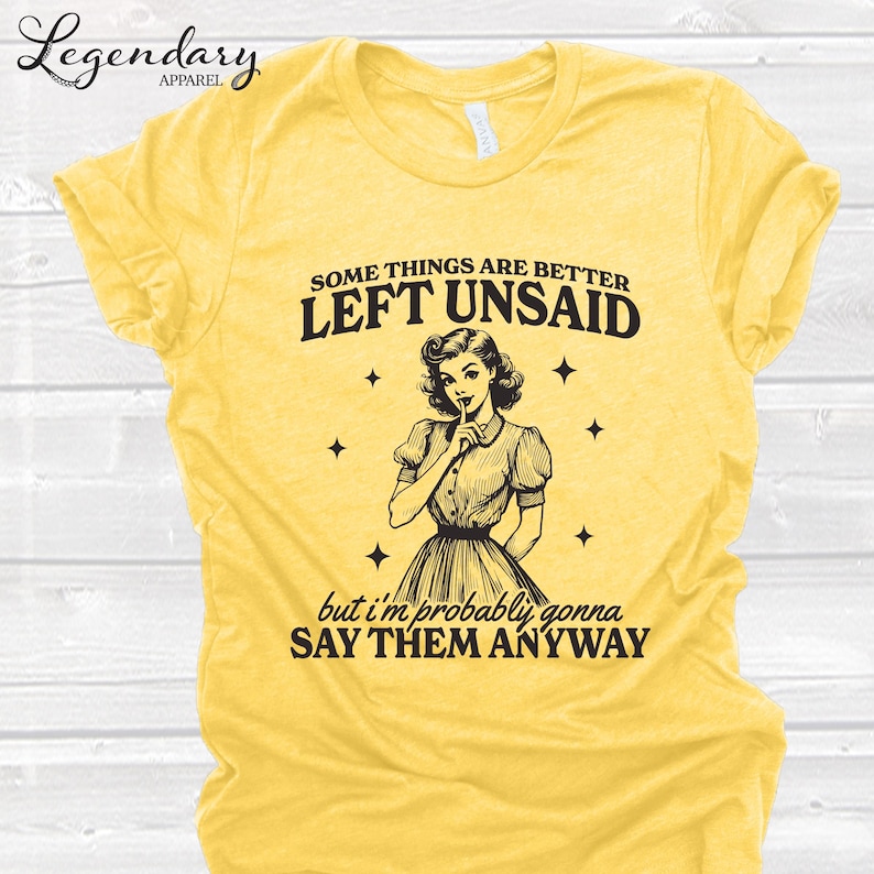Może przedstawiać: Ż&oacute;łta koszulka z napisem "Some things are better left unsaid" i "but I'm probably gonna say them anyway". Koszulka przedstawia ilustrację w stylu vintage kobiety z palcem na ustach. Logo Legendary Apparel znajduje się w lewym g&oacute;rnym rogu.
