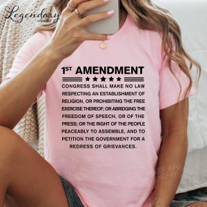 a woman wearing a pink t-shirt with a text overlay that reads "1st Amendment" and " Congress shall make no law respecting an establishment of religion, or prohibiting the free exercise thereof, or abridging the freedom of speech, or of the press, or the right of the people peacefully to assemble, and to petition the government for a redress of Grievances."