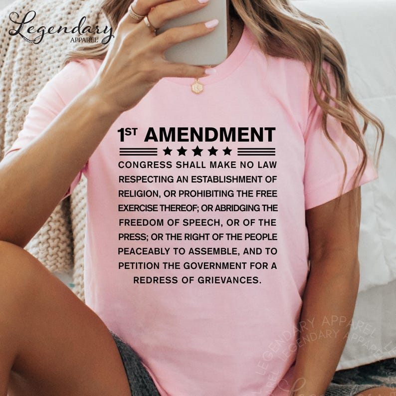 a woman wearing a pink t-shirt with a text overlay that reads "1st Amendment" and " Congress shall make no law respecting an establishment of religion, or prohibiting the free exercise thereof, or abridging the freedom of speech, or of the press, or the right of the people peacefully to assemble, and to petition the government for a redress of Grievances."