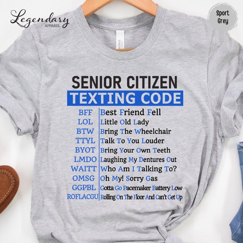 May include: A gray t-shirt with the text "Senior Citizen Texting Code" in blue letters. The shirt lists common texting abbreviations and their meanings for older adults, such as BFF, LOL, BTW, TTYL, BYOT, LMDO, WAITT, OMSG, GGPBL, and ROFLACGU.
