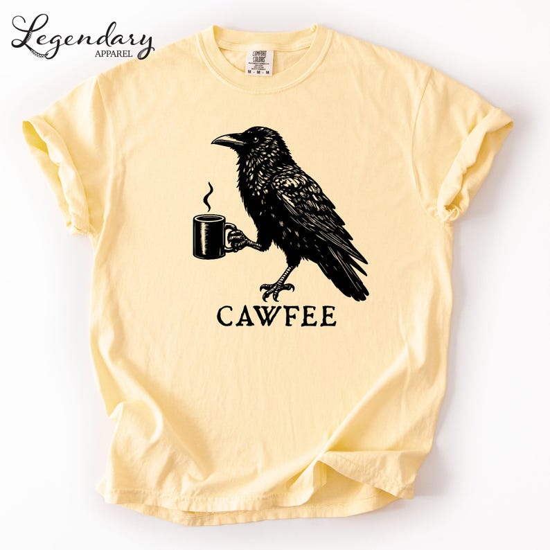 A yellow t-shirt with a black illustration of a crow holding a coffee cup, accompanied by the text "CAWFEE".
