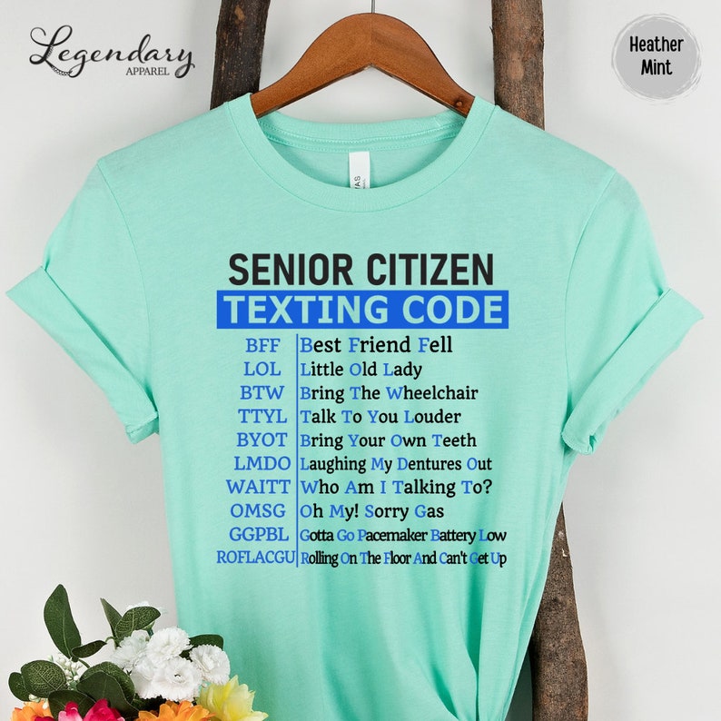 May include: A light blue t-shirt with the text "Senior Citizen Texting Code" in black. The shirt lists common texting abbreviations and their meanings, such as BFF, LOL, BTW, TTYL, BYOT, LMDO, WAITT, OMS, GGPBL, and ROFLACGU.