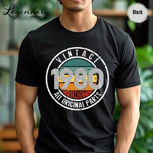 May include: Black t-shirt with a vintage design featuring the year "1980" in a retro font. The design includes the words "VINTAGE" and "ALMOST ALL ORIGINAL PARTS" in a circular layout, with a color scheme of green, orange, and red.