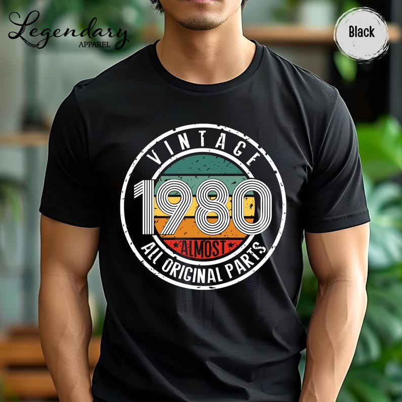 May include: Black t-shirt with a vintage design featuring the year "1980" in a retro font. The design includes the words "VINTAGE" and "ALMOST ALL ORIGINAL PARTS" in a circular layout, with a color scheme of green, orange, and red.