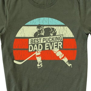 May include: Green t-shirt with a retro-style graphic of a hockey player skating with a hockey stick. The graphic includes the text "Best Pucking Dad Ever".