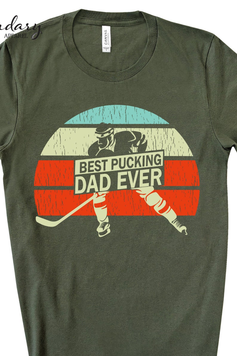 May include: Green t-shirt with a retro-style graphic of a hockey player skating with a hockey stick. The graphic includes the text "Best Pucking Dad Ever".