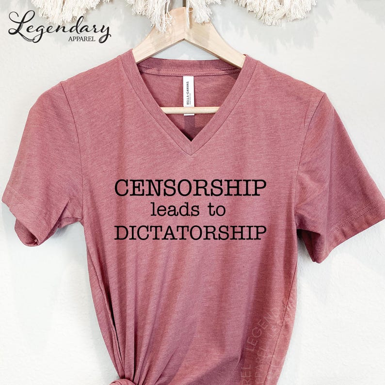 A pink t-shirt with the text "CENSORSHIP LEADS TO DICTATORSHIP" printed on it, hanging on a wooden hanger.