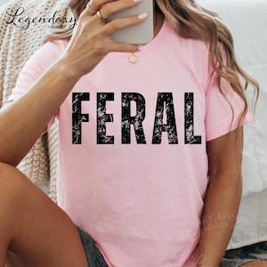 A person is taking a selfie with a pink t-shirt that has the word "FERAL" printed on it.