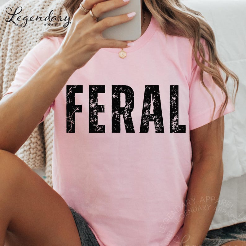 A person is taking a selfie with a pink t-shirt that has the word "FERAL" printed on it.