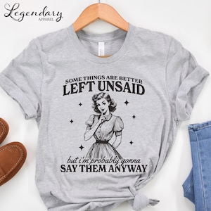 Może przedstawiać: Jasnoszara koszulka z napisem "SOME THINGS ARE BETTER LEFT UNSAID but I'm probably gonna SAY THEM ANYWAY." Koszulka przedstawia ilustrację w stylu vintage kobiety z palcem na ustach.