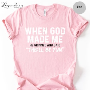 A pink t-shirt with white text that reads "WHEN GOD MADE ME HE GRINNED AND SAID 'THISLL BE FUN'"