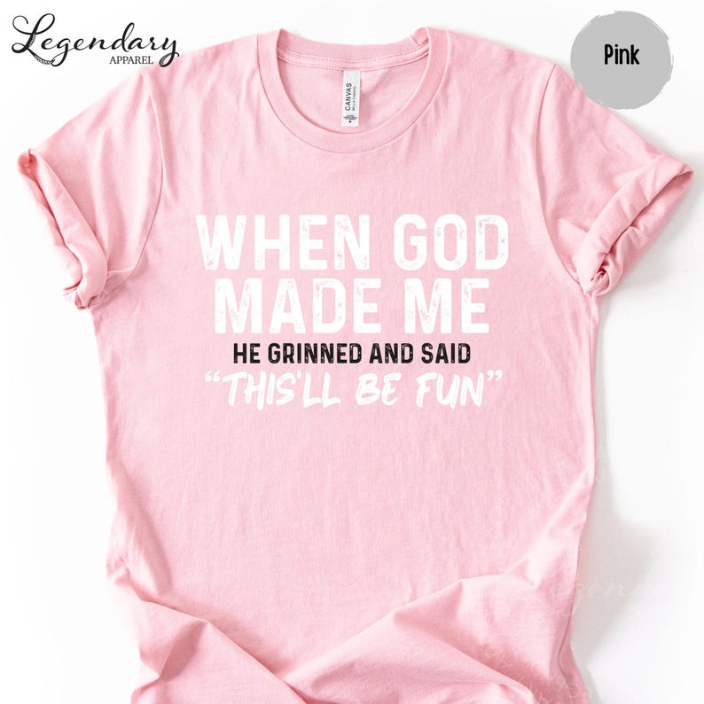 A pink t-shirt with white text that reads "WHEN GOD MADE ME HE GRINNED AND SAID 'THISLL BE FUN'"