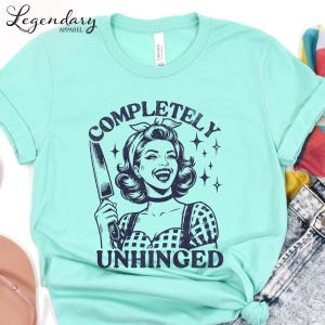 May include: Mint green t-shirt with a graphic of a smiling woman holding a knife, the words "COMPLETELY UNHINGED" are above and below the woman. The shirt has short sleeves and a crew neck.