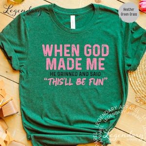 a green t-shirt with a humorous text printed on it, which reads "WHEN GOD MADE ME HE GRINNED AND SAID 'THIS'LL BE FUN'". The t-shirt is displayed on a wooden surface, and there are some other items in the background, such as a pair of shoes and a gold necklace.