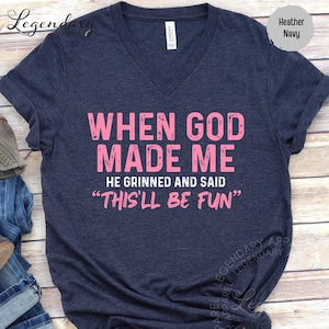 A navy blue t-shirt with a humorous text printed on it, which reads "WHEN GOD MADE ME HE GRINNED AND SAID 'THIS 'LL BE FUN'".