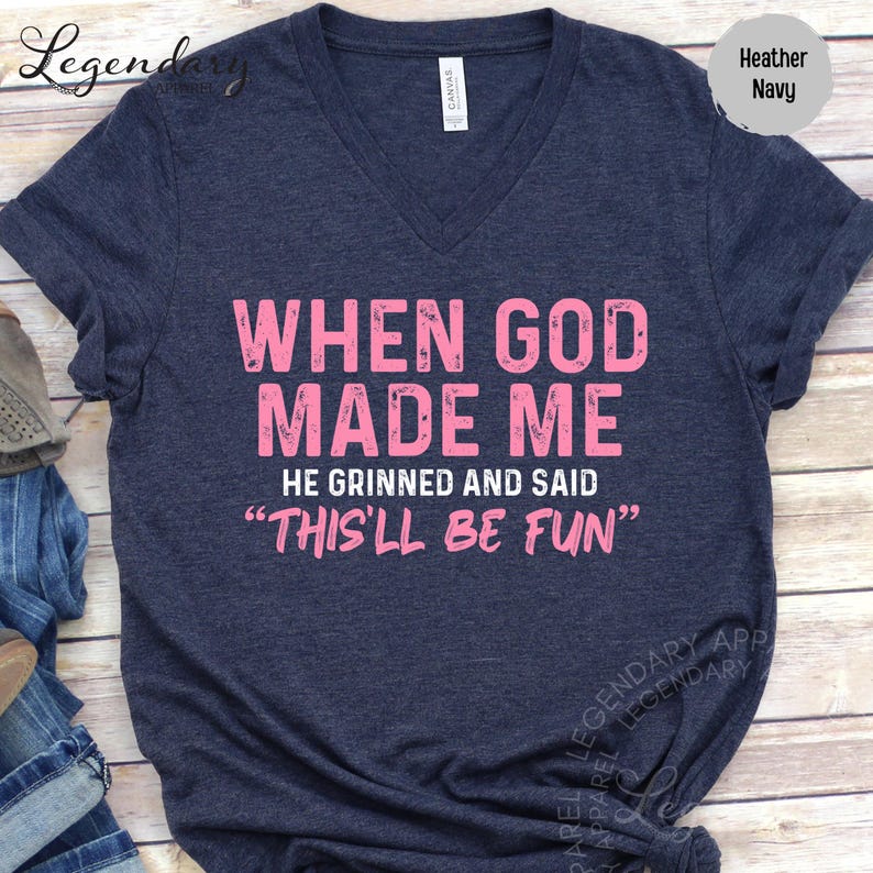 A navy blue t-shirt with a humorous text printed on it, which reads "WHEN GOD MADE ME HE GRINNED AND SAID 'THIS 'LL BE FUN'".