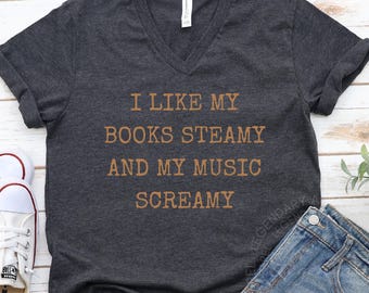 May include: Dark gray V-neck t-shirt with the text "I LIKE MY BOOKS STEAMY AND MY MUSIC SCREAMY" in brown lettering. The shirt is folded, with a pair of white sneakers and blue jeans visible in the background. The Legendary Apparel logo is in the top left.