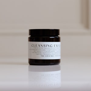 May include: A dark brown glass jar with a black lid containing a cleansing exfoliating scrub. The label reads "Cleansing Exfoliant" and lists ingredients including cane sugar, organic papaya, palm glycerin, charcoal powder, raw honey, pure vitamin E, eucalyptus, and rosemary. The label also includes the brand name "The Coco Bee".