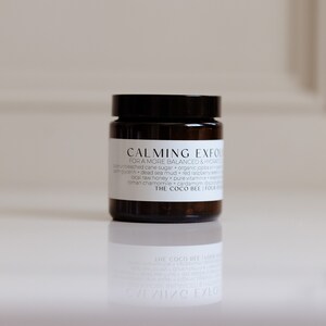 May include: A brown glass jar with a black lid containing a calming exfoliating face mask. The label reads "CALMING EXFOLI  FOR A MORE BALANCED & HYDRATED SKIN" and lists ingredients including cane sugar, organic jojoba oil, dead sea mud, red raspberry, local raw honey, pure vitamin E, and roman chamomile.