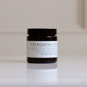 May include: A small, black jar with a white label that reads "Energizing Energy" and "For a more balanced & radiant skin". The label also lists the ingredients: "Unbleached cane sugar + organic avocado oil + palm glycerin + local coffee grounds + local raw honey + cardamom + pure vitamin E + vanilla". The label also includes the text "The Coco Bee Honey" in a smaller font.
