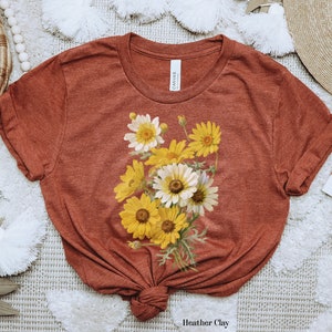 Könnte beinhalten: Ein rostfarbenes T-Shirt mit einem Vintage-Blumendruck aus gelben und weißen Gänseblümchen. Das Shirt ist unten verknotet und hat den Text "Heather Clay" unten aufgedruckt.