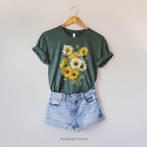 Könnte beinhalten: Ein grünes T-Shirt mit einem floralen Druck aus gelben und weißen Blumen. Das Shirt hängt an einem Holzbügel. Der Text "Heather Forest" ist unter dem Shirt gedruckt.