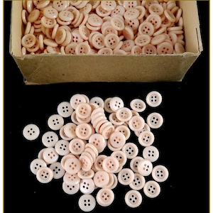 Pode incluir: Uma grande quantidade de botões de plástico rosa claro com quatro furos cada. Os botões estão em uma caixa de papelão e espalhados sobre uma superfície preta.