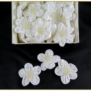 May include: White satin flower embellishments with pearl centers are displayed in a box and scattered on a black surface. The flowers have five petals and are suitable for crafts, scrapbooking, or other decorative projects.