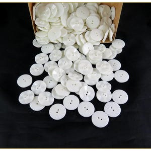 May include: A large collection of round, white, two-hole buttons spilling out from a cardboard box. The buttons are smooth, circular, and feature a slightly concave center with two small holes for sewing, scattered across a dark surface.