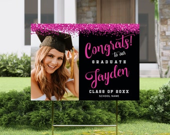 May include: Pink glitter graduation yard sign with a black background. The sign says "Congrats! to our graduate Jayden Class of 20XX School Name". A photo of a graduate in a cap and gown is in the upper left corner.