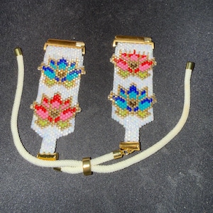 May include: Two beaded bracelets with white backgrounds and gold clasps. Each bracelet features a floral design in pink, blue, and gold beads. The bracelets are connected by a white cord with gold accents.