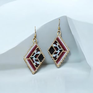 May include: A pair of diamond-shaped beaded earrings. The earrings have gold-coloured hooks and a geometric pattern in black, white, and burgundy. The edges are outlined in gold, creating a contrast. The earrings are displayed on a white surface.