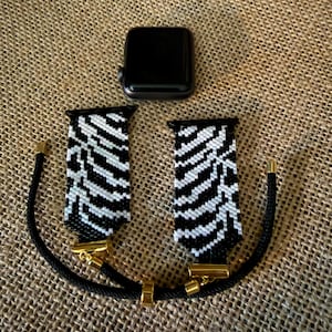 May include: A black smartwatch with a detached two-piece watch band featuring a black and white zebra stripe pattern. The band includes gold-toned metal connectors and a black cord strap with adjustable gold-toned metal beads for a secure fit.