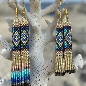 May include: A pair of long, cylindrical beaded earrings featuring a geometric diamond pattern in shades of blue, turquoise, metallic gold, and bronze. The earrings are finished with gold-toned metal hooks and a fringe of dangling metallic gold beads.