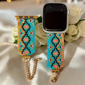 May include: A beaded watch band in turquoise, gold, black, and pink. The band features a geometric pattern and a gold-colored clasp. The watch band is shown with a silver Apple Watch and a separate band with a chain.