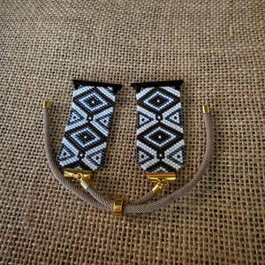 May include: A watch strap replacement featuring two rectangular sections with a black and white geometric diamond pattern. The sections attach to gold-toned metal connectors and are joined by a tan cord with a sliding gold-toned metal bead.