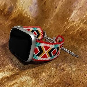 Op de afbeelding: Een zilveren smartwatch met een geweven stoffen horlogeband met een geometrisch patroon in rood, wit, zwart, geel en turkoois. De band is voorzien van een zilverkleurige metalen ketting en zilveren bevestigingsstukken voor de horlogekast.