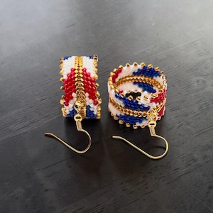 May include: A pair of hoop earrings crafted from small beads in a repeating pattern of red, white, and blue. The beads are arranged in a cylindrical shape with a gold-toned metal wire running through the center. Each earring features a gold-toned hook.