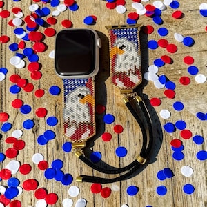 May include: A smartwatch band featuring a beaded eagle design with red, white, and blue colors. The band includes gold-tone metal connectors and a black cord with gold-tone beads for adjustment, attached to a silver-toned smartwatch case.