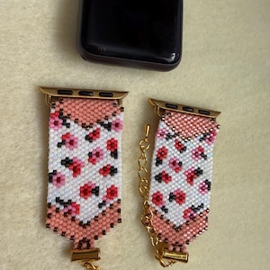 以下が含まれることがあります： ピンク、白、黒の花柄のビーズApple Watchバンドが2つ。 バンドには金色の金具とロブスタークラスプが付いています。 黒いApple Watchが背景にあります。